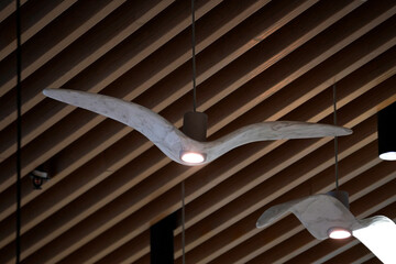 Unique ceiling lights shaped like flying birds hang from a wooden slatted ceiling. Each light emits...