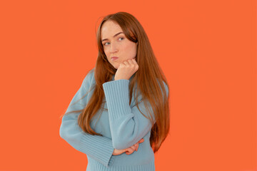 Young woman with long brown hair poses thoughtfully against a bright orange background while...