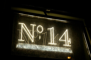 Illuminated vintage-style neon sign displaying "No 114" in white light. The sign's glass surface shows reflections and slight wear, set against a dark background, evoking nostalgic urban ambiance.

