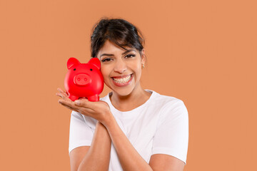 Smiling woman holds red piggy bank against a warm orange background while sharing her enthusiasm...