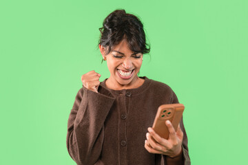 Excited woman celebrates a success while looking at her phone against a bright green background in...