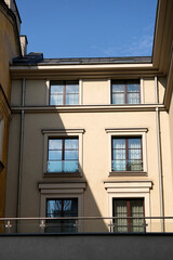 Obraz premium Facade of a beige residential building with symmetrical windows reflecting blue sky. Modern glass balcony railing contrasts with classic architectural details. Bright sunlight highlights clean design.