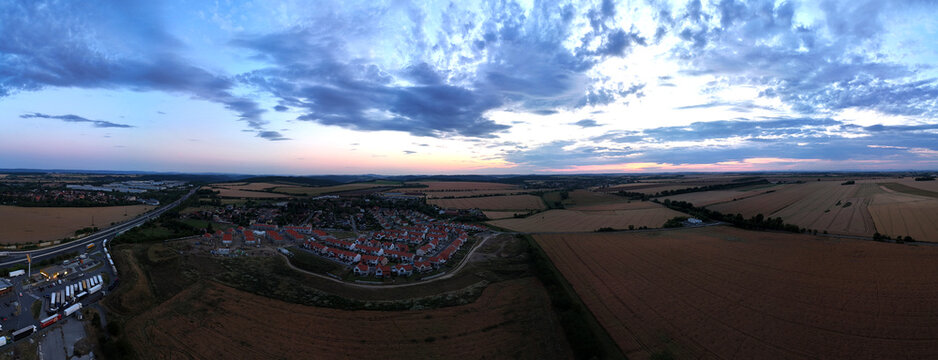 Panoramic aerial shot of a rural landscape at dusk. The sun is setting behind distant hills, casting a warm glow on the horizon under a sky filled with dramatic clouds. Below, farmland and a small vil