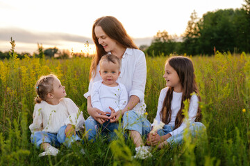 Fototapeta premium A joyful mother sits in a sunlit grassy field surrounded by her three children (two girls and a baby), all wearing white tops and denim, enjoying the outdoor evening.