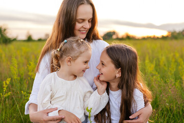 A happy mother embraces her two young daughters in a golden field at sunset, as the girls lovingly...