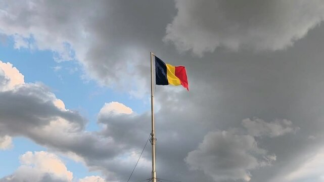 The national flag of Romania waves proudly on a tall pole under a dramatic sky with contrasting clouds and patches of blue.