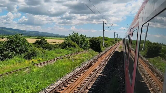 A red passenger train moves along a double-track railway through wide rural fields under a partly cloudy summer sky , in Romania
