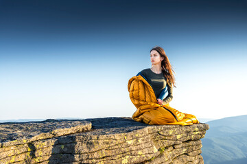 Woman sitting wrapped in a yellow sleeping bag on mountain cliff, gazing into distance during sunset