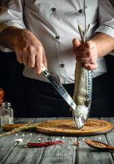 Obraz premium A chef holds a freshly caught fish with one hand while expertly filleting it with a knife in a cozy kitchen. Various spices and herbs surround the wooden cutting board, creating a vibrant atmosphere