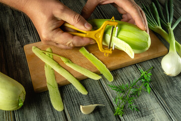Hands are peeling zucchini on a wooden cutting board, surrounded by fresh vegetables like spring...