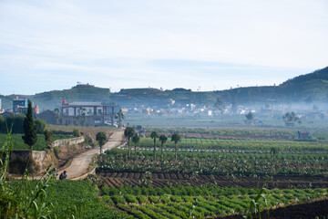 A wide shot of a rural Indonesian village nestled at the foot of a lush green mountain, partially...