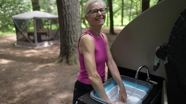 Portrait of retired woman happily washes dishes at a campground, surrounded by trees, with a cozy teardrop camper trailer in the background, embracing the camping lifestyle.