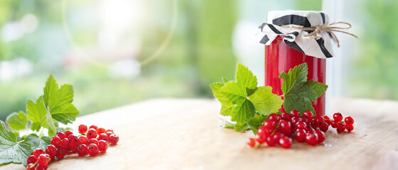 Fresh homemade red currant jam in jar with currants and green leaves on wooden table against light bokeh background. Horizontal close-up with space for text.