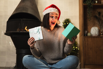 Merry Christmas. Young woman in Santa hat unpacking open up gift box near Christmas tree. Girl in...
