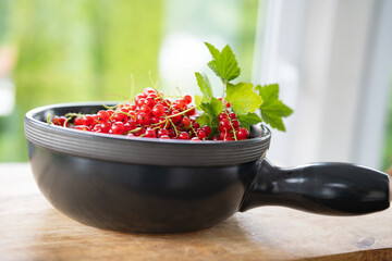 Freshly picked ripe red currants in a dark ceramic bowl, decorated with green leaf on wooden table against bright background. Close-up of healthy seasonal fruits.