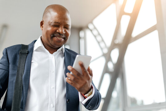Smiling businessman looking at phone screen in bright airport space. His relaxed stance and modern look reflect confidence and digital connectivity while traveling, copy space