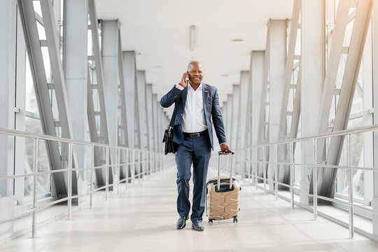 Smiling African American businessman talking on cellphone while walking confidently through a modern airport terminal, black male pulling suitcase and carrying a leather briefcase