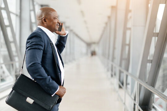 Smiling black businessman in suit talking on cellphone in airport, side view shot of african american entrepreneur having mobile call while waiting flight at terminal, copy space
