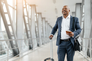 Black businessman with suitcase and passport in hand standing confidently in bright airport...