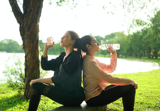 Thirsty young woman and her friends are drinking water. Hot day. Young woman, jogging, playing sports and living a healthy life. Refreshing. Hot and humid summer weather in the park.