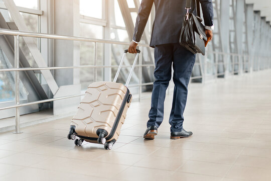Business traveler walking through airport terminal pulling suitcase with one hand, briefcase in the other. Confident posture and sharp suit reflect executive energy, cropped