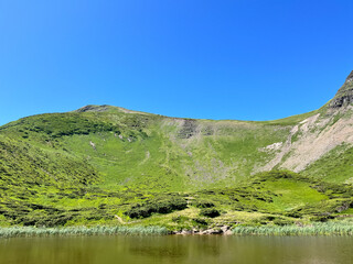 Lush green mountain under a clear blue sky by a calm water body.