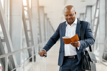 Black businessman holding suitcase and reading travel documents at airport. Focused expression,...