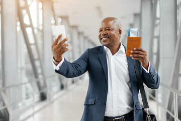 Happy businessman taking selfie with smartphone in airport while holding passport. Bright terminal...