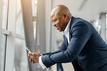 Black businessman leaning on airport railing smiling while reading something on smartphone. Calm...