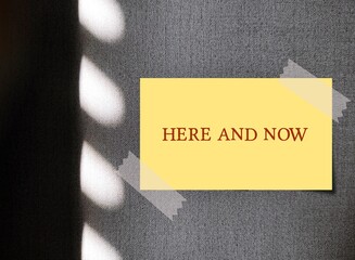 Yellow note on wall with text written HERE AND NOW - refers to present moment, being conscious and...