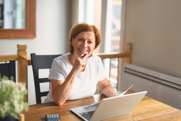 happy senior woman calculating family budget, sitting at kitchen counter with laptop, papers,