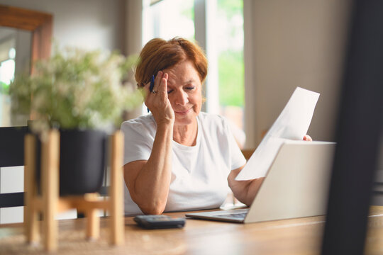 Sad frustrated senior woman pensioner having depressed look, holding hand on her face, calculating family budget, sitting at kitchen counter with laptop, papers, coffee, calculator and cell phone - Powered by Adobe