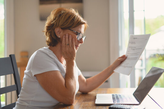 Sad frustrated senior woman pensioner having depressed look, holding hand on her face, calculating family budget, sitting at kitchen counter with laptop, papers, coffee, calculator and cell phone