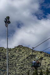 Two people ride a mountain chairlift over a rocky slope under a partly cloudy sky. High-altitude...