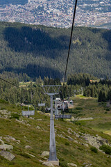 
A view from a mountain chairlift descending over a rocky, forested slope with a city sprawling in...