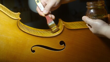 Skilled luthier applying varnish on cello body, holding stained jar, highlighting intricate...