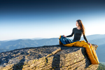 Young woman camping in mountains, sitting in sleeping bag on cliff at sunrise, enjoying peaceful moment