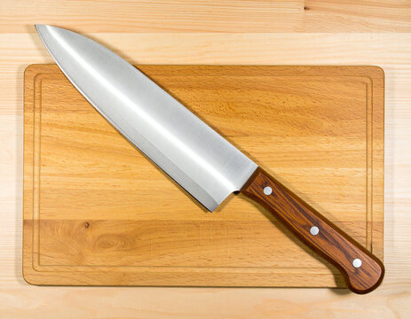 A large, gleaming chef's knife with a brown wooden handle lies diagonally on a clean, light-colored wooden cutting board, ready for food preparation. The board has a subtle juice groove around its per