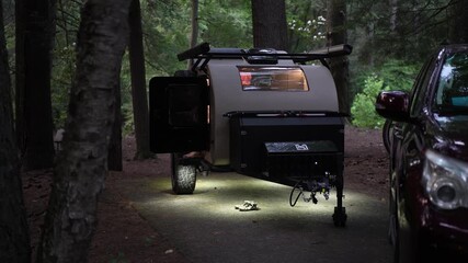 Under the night sky, a mature woman steps out of her teardrop trailer in a tranquil forest setting, embracing an adventurous camping lifestyle.