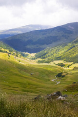 Grassy mountain valley with scattered rocks and a winding stream flows beneath partly cloudy skies....