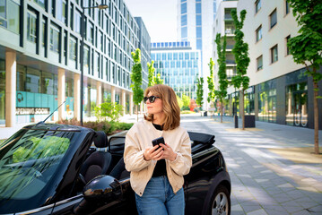 A middle aged woman standing next to her car using a smartphone