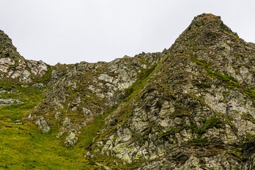 Rock-strewn green slopes ascend sharply toward jagged mountain ridges beneath an overcast sky. The...