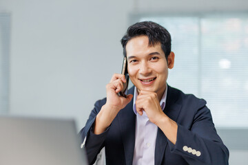 Portrait of happy manager speaking on mobile phone and looking at camera while working on laptop in...