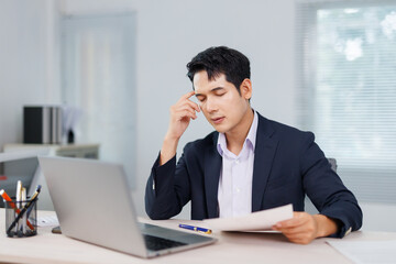Naklejka premium Young Asian manager experiencing a headache while working with documents and a laptop in a busy office environment, feeling overwhelmed