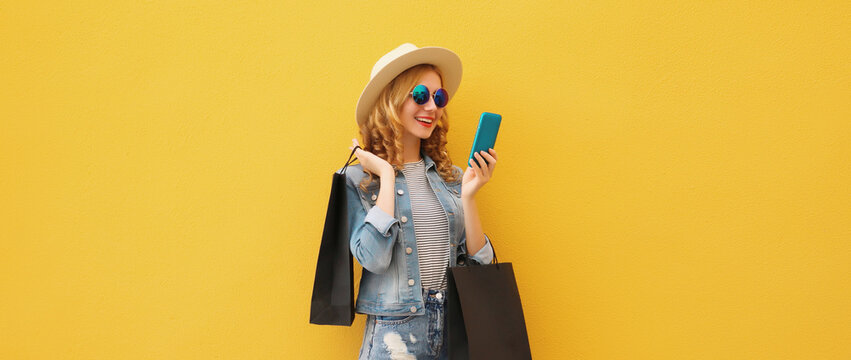 Beautiful happy young woman holding phone with black shopping bag, summer hat on yellow background