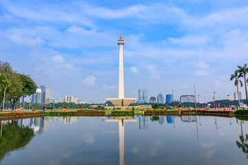 National Monument (Monas) is iconic landmark in the center of Jakarta reflected in a calm pool with palm trees and blue sky.