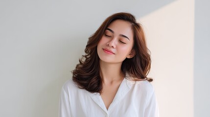 Obraz premium Young woman with long wavy hair smiling softly in natural light, wearing a white shirt, embodying joy and tranquility in a minimalist space