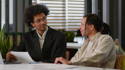 Two multiracial business men African American businessmen partners diverse colleagues males discuss financial data analyzing documents marketing project result report use laptop talk at office desk