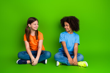 Two cheerful young girls in colorful outfits sitting cross-legged on a bright green background...
