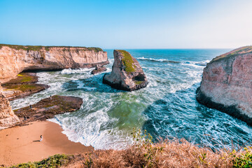 Iconic Shark Fin Cove near Davenport, California, captured from above. Dramatic rock formations, sea cave, and ocean waves meet the rugged coastline under a bright blue sky